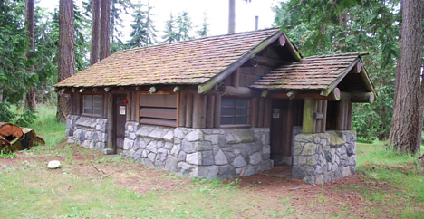 cabin in grassy woods. Top half is made of logs and a wooden roof. Bottom half is made of stone.