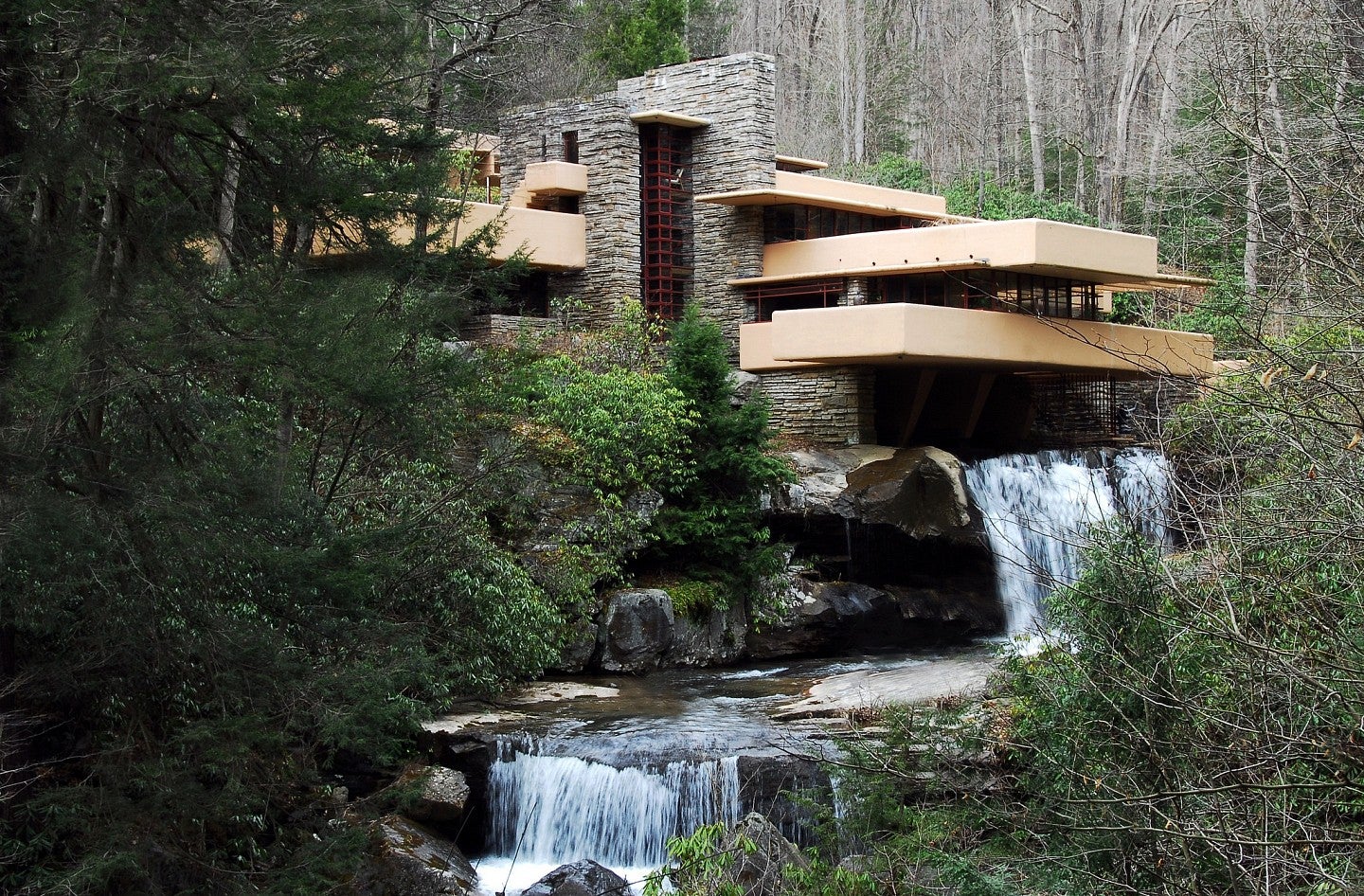 Color photo of the iconic Fallingwater. Shows a modern design house over a waterfall in a scenic forest scape. 