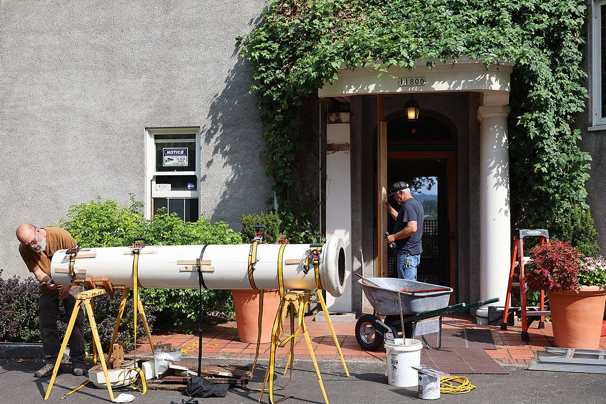 Preservationists work on a column outside the historic garden house. Shows a tall, white column strapped horizontally so a preservationist can work on the interior of the column. 