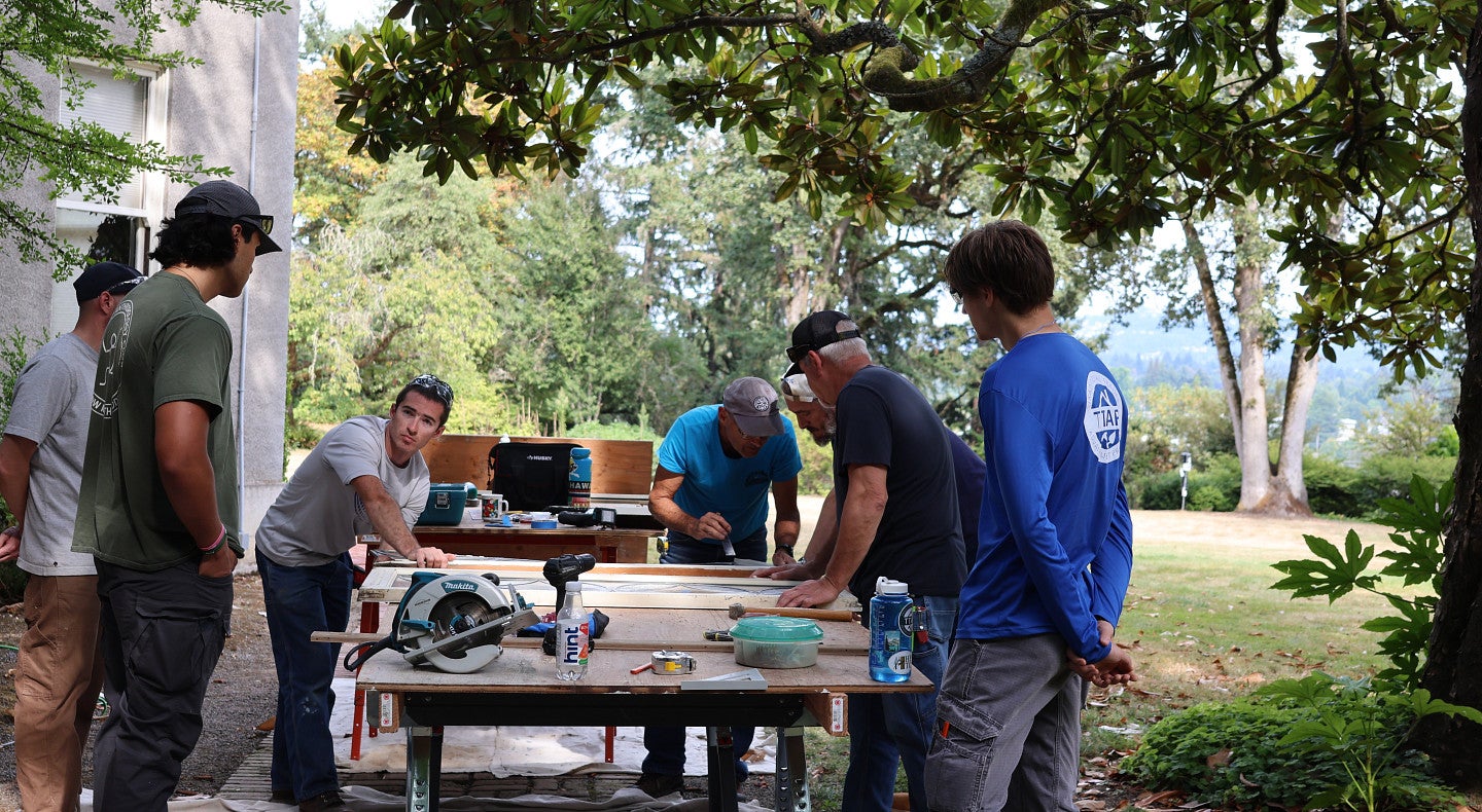 Students and preservationists work on windows outdoors in Portland surrounded by beautiful gardens and trees.