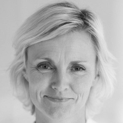 Photograph of Tine. Shows a women in front of a white wall with light colored, short, neck length hair.